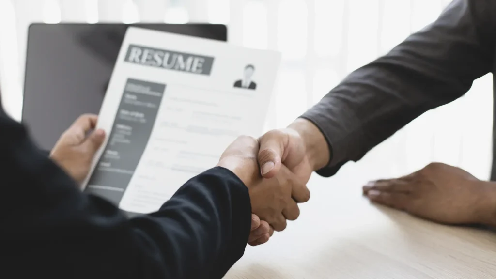 Two people shaking hands with a resume visible in the background.